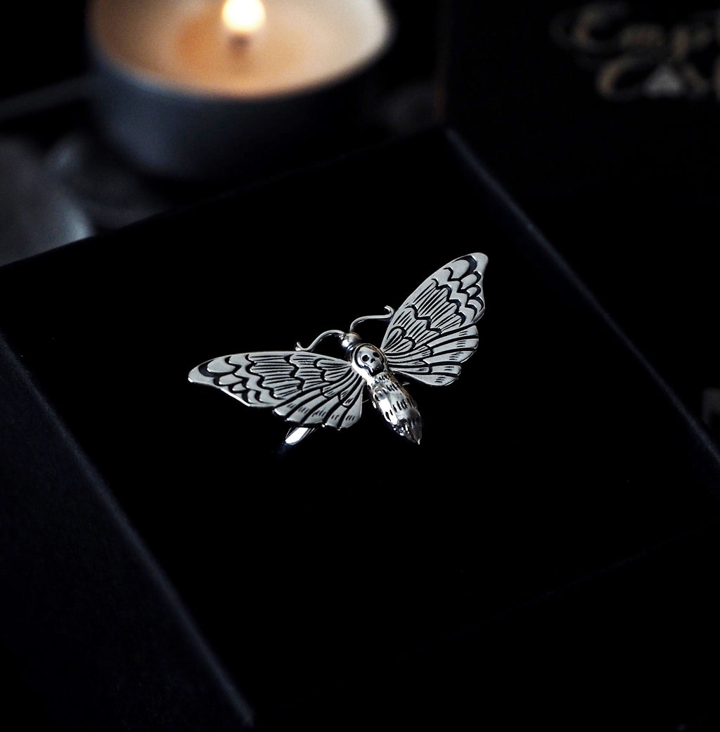 a picture of a sterling silver death's head hawkmoth ring set in a black ring box infront of a glowing candle. 